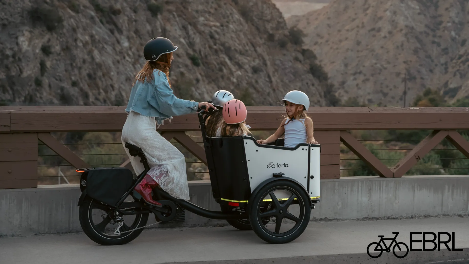 Woman riding a Ferla Royce MAX cargo trike with three children across a bridge at sunset